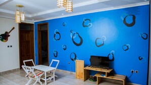 A room with a blue wall, a wooden TV stand, a white table with chairs, and a guitar on the wall. - Green Eskapes Suites (Kigali)