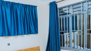 A bedroom with a wooden bed, white pillows, a floral bedspread, blue curtains, and a window with bars. - Green Eskapes Suites (Kigali)