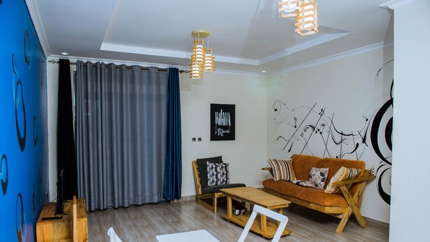 A modern living room with a sofa, a coffee table, and a white table. There is a blue wall with abstract designs and a wooden floor. - Green Eskapes Suites (Kigali)