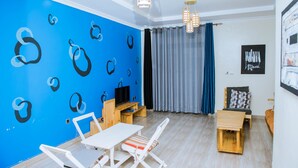 A modern living room with a blue wall, wooden furniture, a TV, and a window with curtains. - Green Eskapes Suites (Kigali)