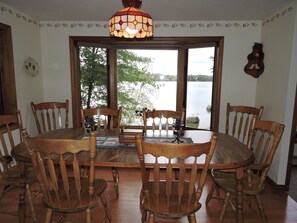 Un coin repas avec une table ronde en bois et des chaises, un luminaire en vitrail et une vue sur un lac, visible à travers de grandes fenêtres.