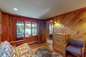 Interno di una baita con pavimento in legno, un divano, una poltrona, un comò e una porta che dà su un balcone con vista sugli alberi.