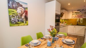 A dining area with a wooden table set for four, green chairs, and a painting of sheep on the wall. - Herdwick Cottage (Brampton)