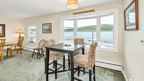 Een eetgedeelte met een tafel en stoelen, een raam met uitzicht op een meer, en een bord boven het raam waarop staat: 'Life is good at the lake'.
