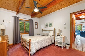 Una camera da letto con soffitto in legno, un letto con testiera bianca e dorata, un comodino, una cassettiera e una finestra con tende.