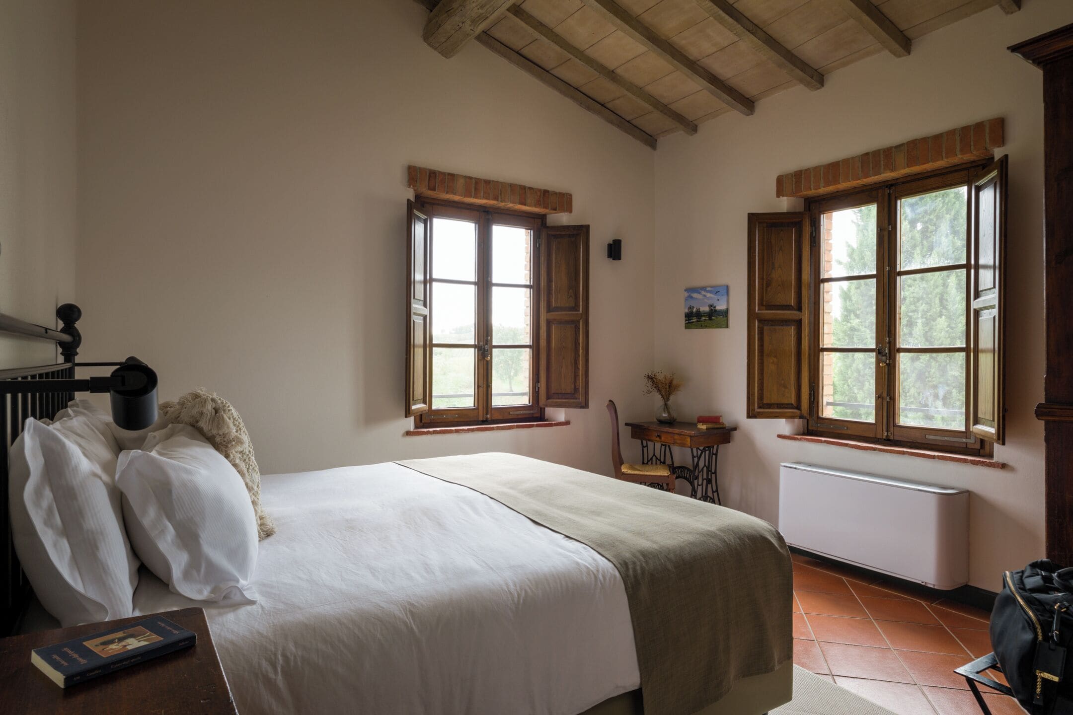 Un dormitorio con cama, mesita de noche, silla, radiador y dos ventanas con persianas de madera.