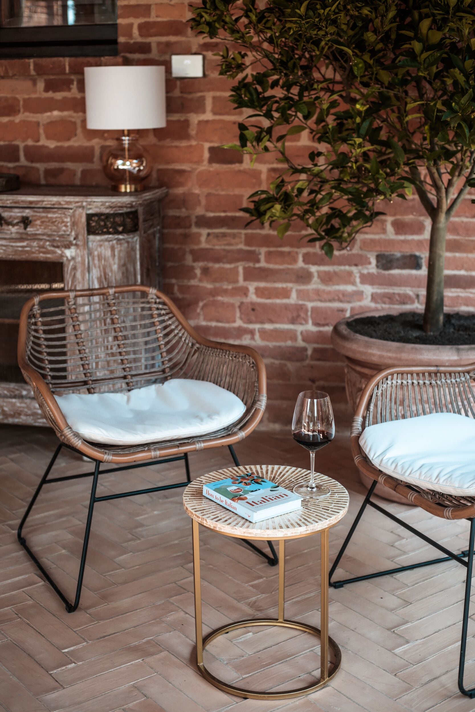 A cozy seating area with wicker chairs and a small round table, a potted plant, and a lamp against a brick wall.