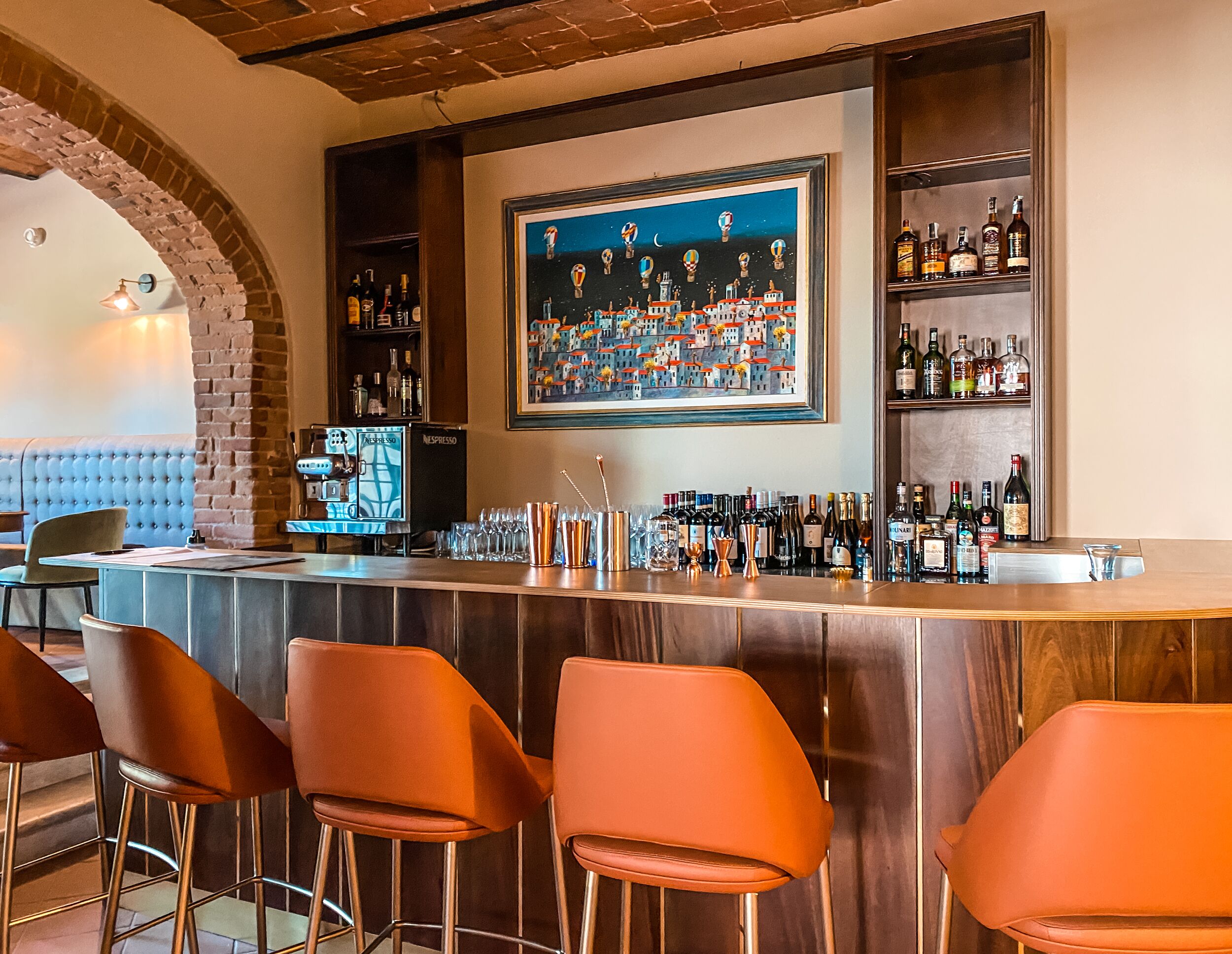 A bar area with a wooden counter, orange bar stools, a Nespresso machine, and a large framed painting of hot air balloons.