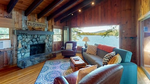 A cozy cabin interior with a stone fireplace, wooden walls, a sofa, and a view of a lake. - Pet Friendly waterfront house on Lake Winnipesaukee sleeps 6 (Moultonborough)