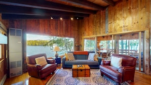 A cozy living room with a wooden ceiling, a sofa set, a patterned rug, and a large window offering a view of trees and a body of water. - Pet Friendly waterfront house on Lake Winnipesaukee sleeps 6 (Moultonborough)