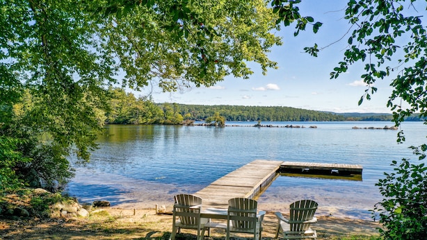A lakeside dock with two Adirondack chairs and a table, surrounded by trees and a clear view of the lake. - Pet Friendly waterfront house on Lake Winnipesaukee sleeps 6 (Moultonborough)