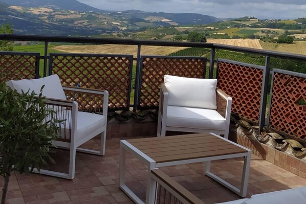 Un balcon avec deux chaises blanches et une table en bois, donnant sur un paysage pittoresque composé de champs et de collines.