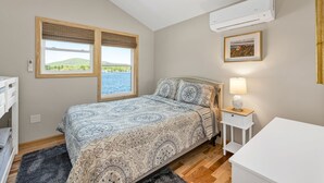 Ein Schlafzimmer mit Bett, Nachttisch mit Lampe, Fenster mit Blick auf Grünflächen und Wasser sowie eine Klimaanlage an der Wand.