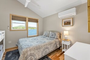 Une chambre à coucher avec un lit, une table de chevet avec une lampe, une fenêtre donnant sur un paysage verdoyant et une étendue d’eau, et un climatiseur fixé au mur.