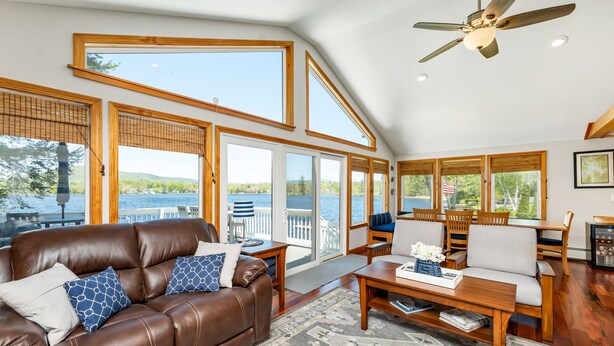 Amplia sala de estar con un sofá de cuero marrón, una mesa de centro de madera y ventanas grandes que ofrecen vistas a un lago.