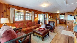 Un intérieur de chalet chaleureux, avec un revêtement en bois, un canapé, un fauteuil, un coin repas et une cuisine.