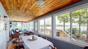 Un espace repas abrité avec une table dressée pour quatre personnes, de grandes fenêtres offrant une vue sur l’extérieur et un plafond en bois.