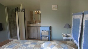 Ein Schlafzimmer mit Bett, Nachttisch, Lampe sowie einem Badezimmer mit Dusche, Waschbecken und Spiegel.