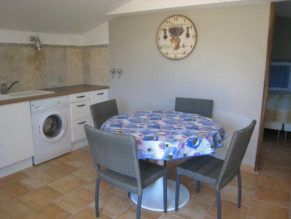 A compact kitchen with a washing machine, a dining table with four chairs, and a wall clock. - COMFORTABLE GITE SWIMMING POOL GARDEN (Forcalquier)