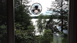 Een raam met een glas-in-lood decoratie van een vogel, omringd door weelderige begroeiing en uitzicht op een meer.