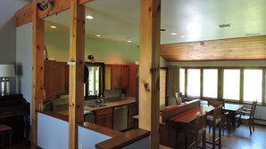 Cozinha moderna com vigas de madeira, área de refeições com várias cadeiras e grandes janelas que proporcionam luz natural.