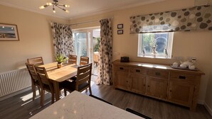 A dining area with a marble table and wooden chairs, a wooden cabinet with kitchenware, and a window with curtains. - The Emberton (Bridlington)