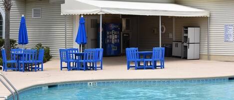 A poolside area with blue chairs and umbrellas, a Pepsi vending machine, and a covered seating area.