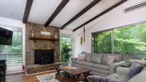 A living room with a stone fireplace, a gray sofa, a wooden coffee table, and a large window with a view of greenery. - The Sunset Chalet | Pet-friendly, Deck & 5 Minutes to Black Mountain! (Black Mountain)