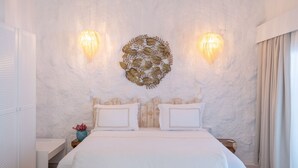 Un dormitorio con cama blanca, dos apliques de pared, una obra de arte con motivos de hojas y una ventana con cortinas.