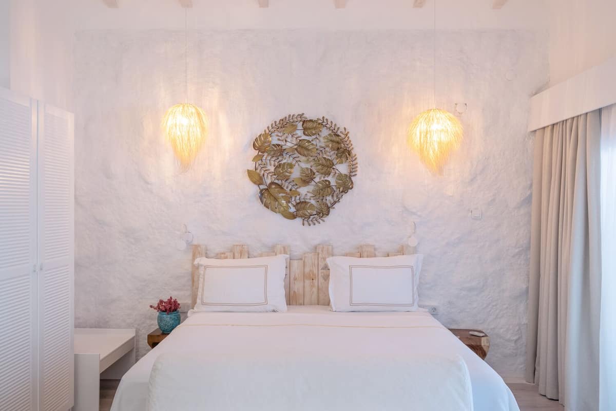 Un dormitorio con cama blanca, dos apliques de pared, una obra de arte con motivos de hojas y una ventana con cortinas.