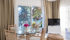 Una habitación con una mesa de vidrio rodeada de sillas de mimbre, un televisor sobre un mueble blanco, y vistas a árboles y agua desde el exterior.