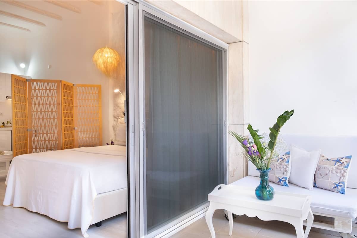 Habitación de hotel con una cama, una puerta corrediza de vidrio, un sofá blanco y una pequeña mesa con un florero.