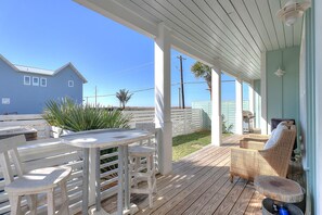 En overdækket udendørs terrasse med træterrasse, hvide møbler og udsigt til et hus og palmer.