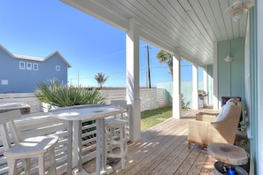 En overdækket udendørs terrasse med træterrasse, hvide møbler og udsigt til et hus og palmer.
