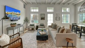 A spacious living room with a large TV, comfortable seating, and multiple windows providing natural light.