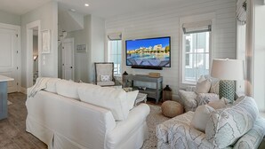 A living room with a large flat-screen TV, white sofas, patterned cushions, and a ceiling fan. - Surf & Sand 235 LAK (Port Aransas)