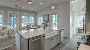 A modern kitchen with a central island, stainless steel appliances, pendant lighting, and large windows. - Surf & Sand 235 LAK (Port Aransas)