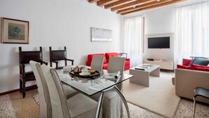 Una sala de estar moderna con una mesa de comedor de vidrio, sofás rojos y color beige, sillas de madera, un televisor de pantalla plana y obras de arte en las paredes.