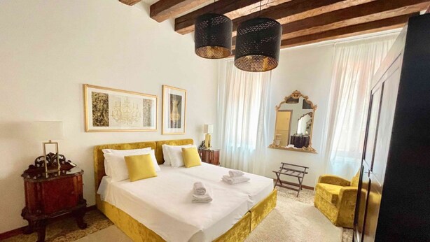 Una habitación de hotel con una cama grande, un cabecero de madera, un espejo, un sillón amarillo y una araña.