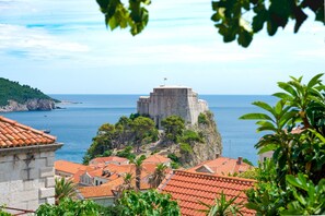 Un pueblo costero con una imponente fortaleza de piedra que domina el mar, rodeado de edificios con tejados de tejas rojas y abundante vegetación.