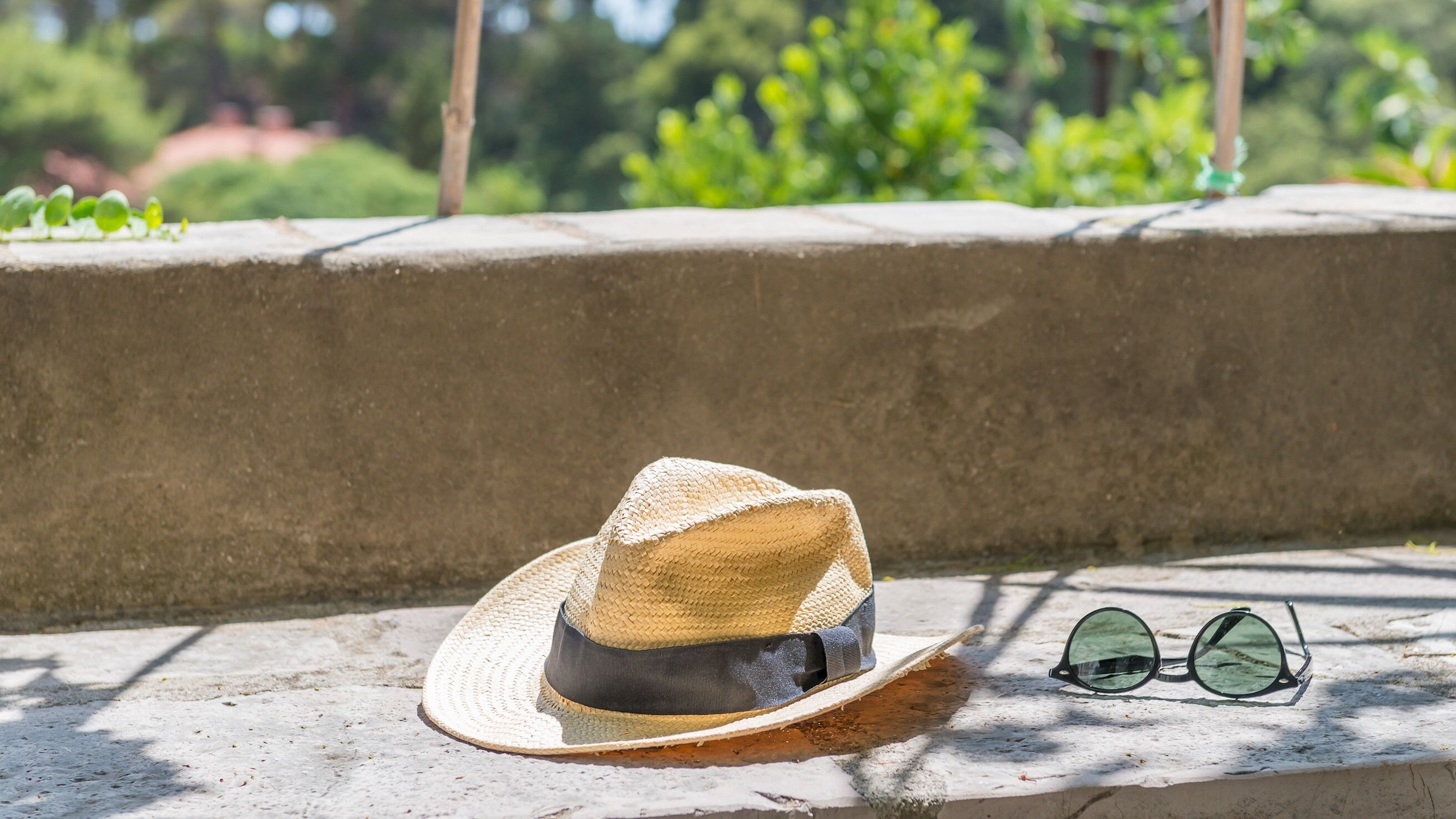 A straw hat and sunglasses on a concrete ledge with greenery in the background.