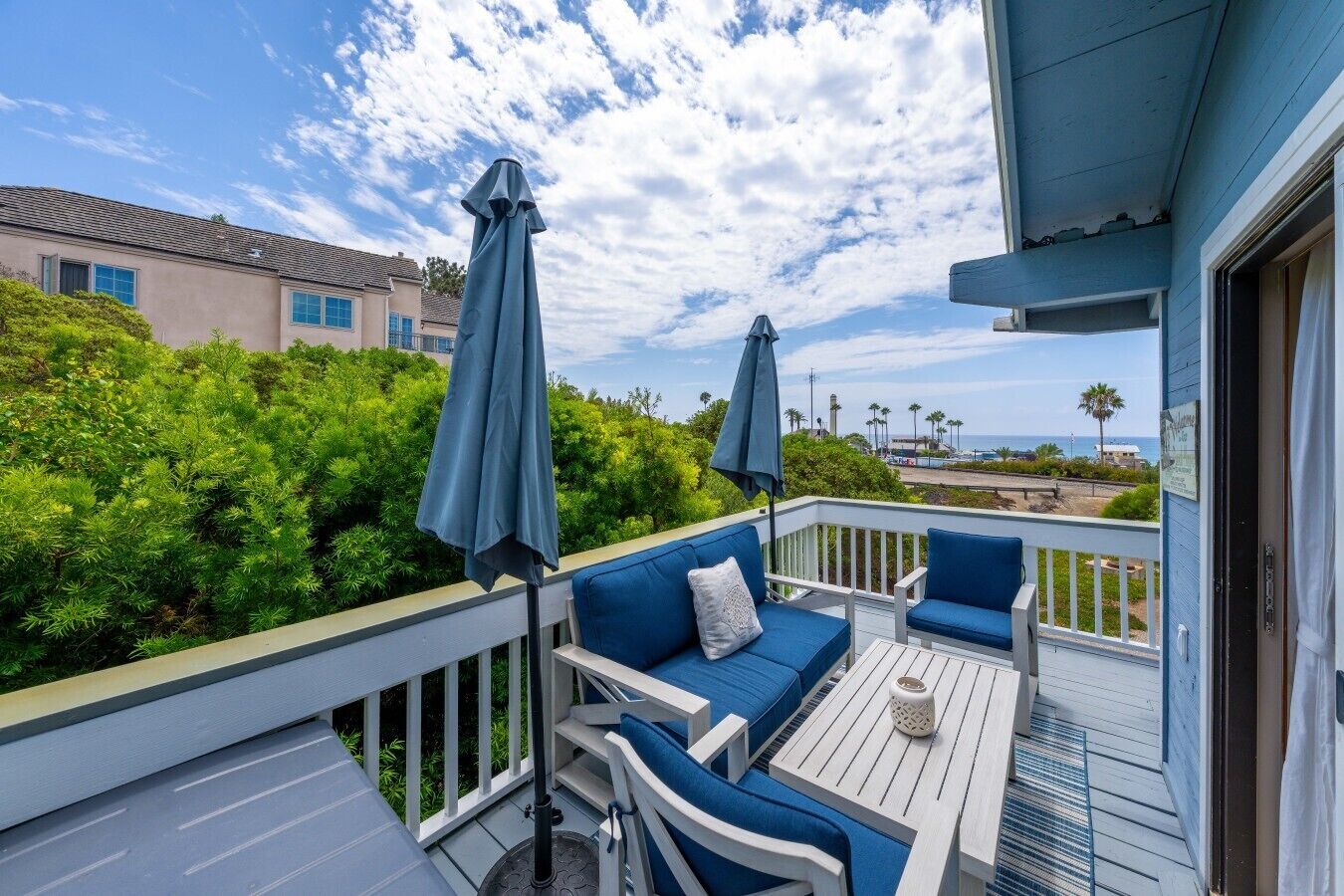 A balcony with blue outdoor furniture, two umbrellas, and a view of the ocean and palm trees.