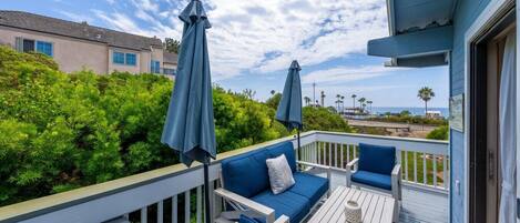 A balcony with blue outdoor furniture, two umbrellas, and a view of the ocean and palm trees.