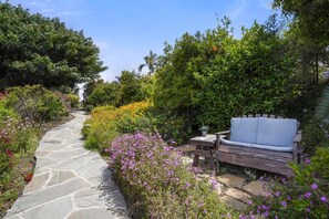 A stone pathway leading to a wooden bench with a table, surrounded by lush greenery and flowering plants.
