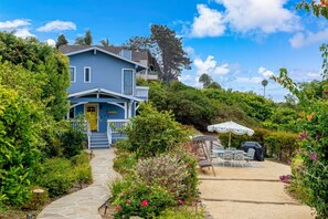 A two-story house with a front porch, a garden with various plants and flowers, and outdoor furniture. - Darling Beach Cottage, Ocean Views, central (Del Mar)