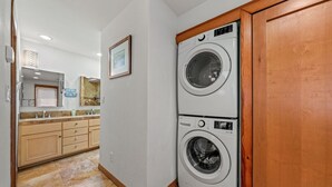 A laundry room with two stacked washing machines, a wooden cabinet, and a kitchen area in the background. - Darling Beach Cottage, Ocean Views, central (Del Mar)