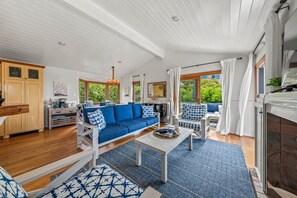 A spacious living area with a blue sofa, a coffee table, and a view of the outdoors through large windows. - Darling Beach Cottage, Ocean Views, central (Del Mar)