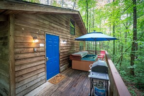 A wooden cabin with a blue door, a hot tub, and a barbecue grill on the deck surrounded by trees.