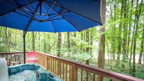 A hot tub on a wooden deck with a blue umbrella overhead, surrounded by a forested area.