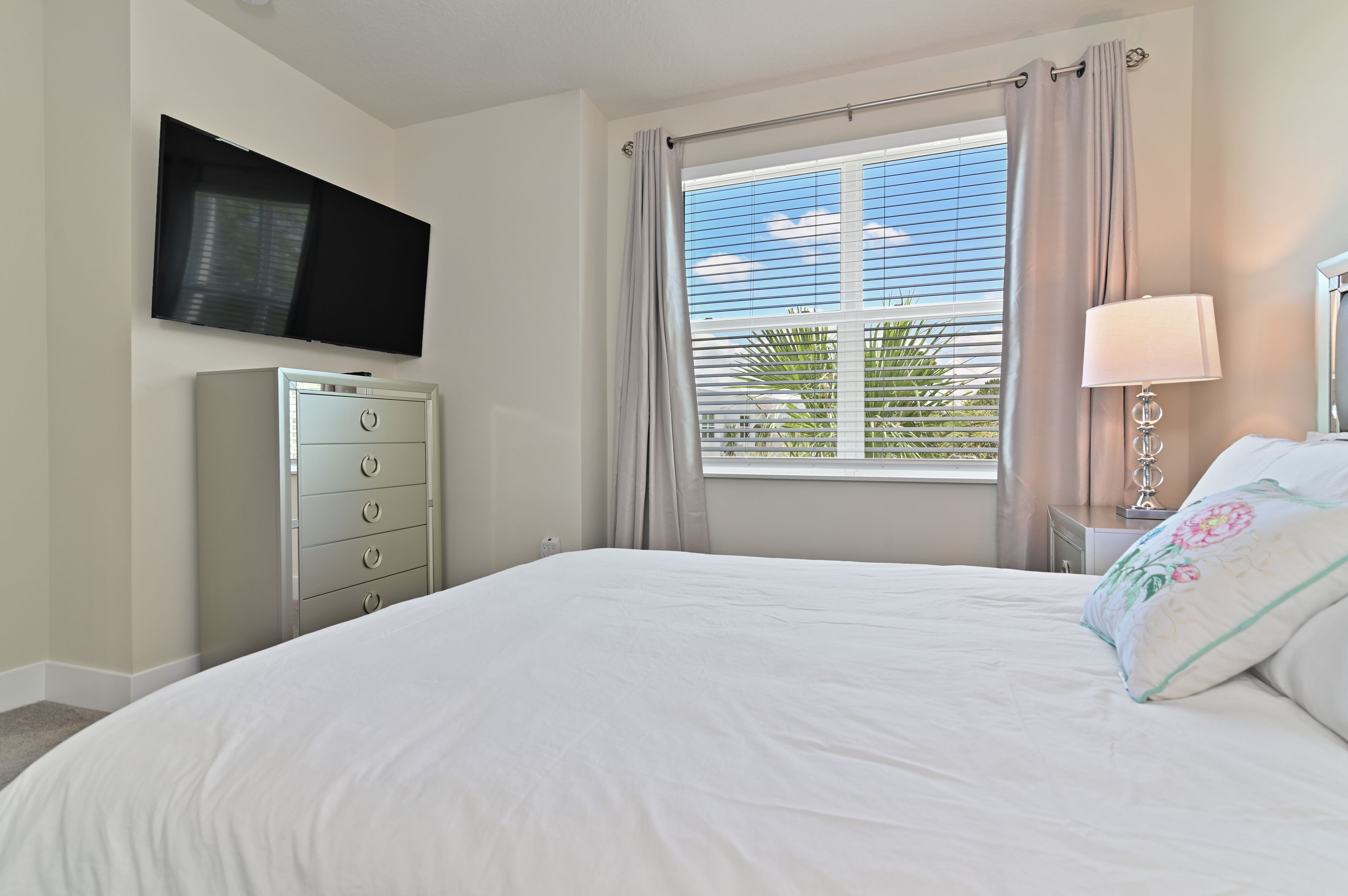 Un dormitorio con cama, televisor montado en la pared, cómoda, ventana con cortinas y mesita de noche con lámpara.